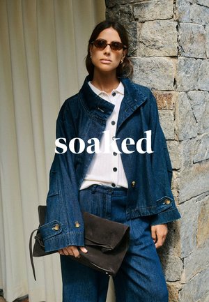 Jeansjacke mit lockerem Schnitt, goldfarbenen Knöpfen und weiten Ärmeln, kombiniert mit einem weißen Hemd und einer dunklen Wildleder-Clutch.