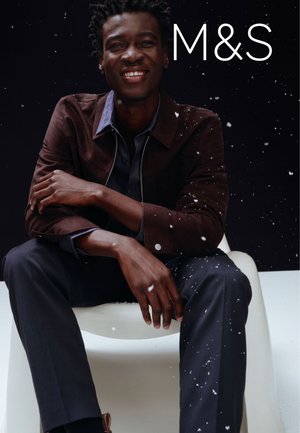 Bruine jas met ritssluiting over een donker shirt; zittende man tegen een donkere achtergrond met sneeuwvlokken. Donkere pantalon; minimalistische stoel. M&S-logo.