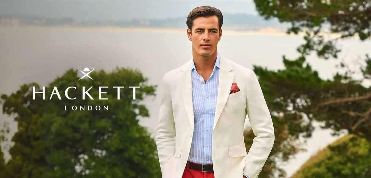 Hombre con blazer blanco, camisa azul a rayas y pantalones rojos está de pie al aire libre con árboles y agua al fondo.
