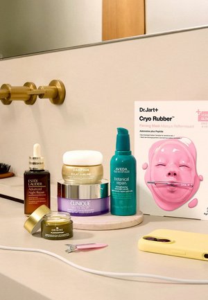 Cinque prodotti per la cura della pelle tra cui un siero Estée Lauder, una crema Darphin, un balsamo Clinique, un siero Aveda, una crema Origins, un tagliaunghie rosa e una custodia per telefono gialla su un piano del bagno.
