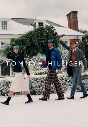 Tre persone camminano nella neve portando un albero di Natale. Sono vestite con abiti invernali: giacca imbottita blu, blazer nero e pantaloni a quadri.