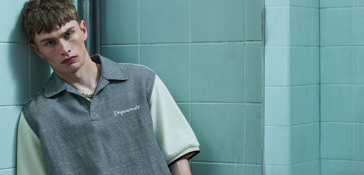 Joven con el pelo corto apoyado contra una pared de azulejos verde azulado, vistiendo una camisa de manga corta a cuadros con mangas blancas.