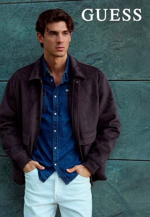 Chaqueta de ante marrón sobre una camisa vaquera, combinada con pantalones blancos. Modelo posa con las manos en los bolsillos frente a un fondo de piedra verde.