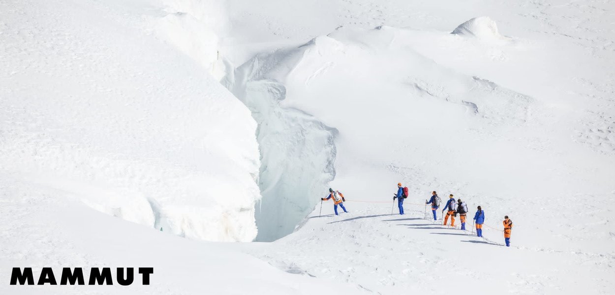 Sieben Bergsteiger in blauer und oranger Ausrüstung sind zusammen am Seil gesichert und wandern über einen schneebedeckten Gletscher in der Nähe einer tiefen Gletscherspalte, im Eck das Mammut-Logo.