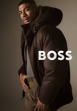 Veste rembourrée marron foncé avec grande capuche, tissu texturé et détails à cordon de serrage. Portée par-dessus un haut gris et un pantalon kaki.