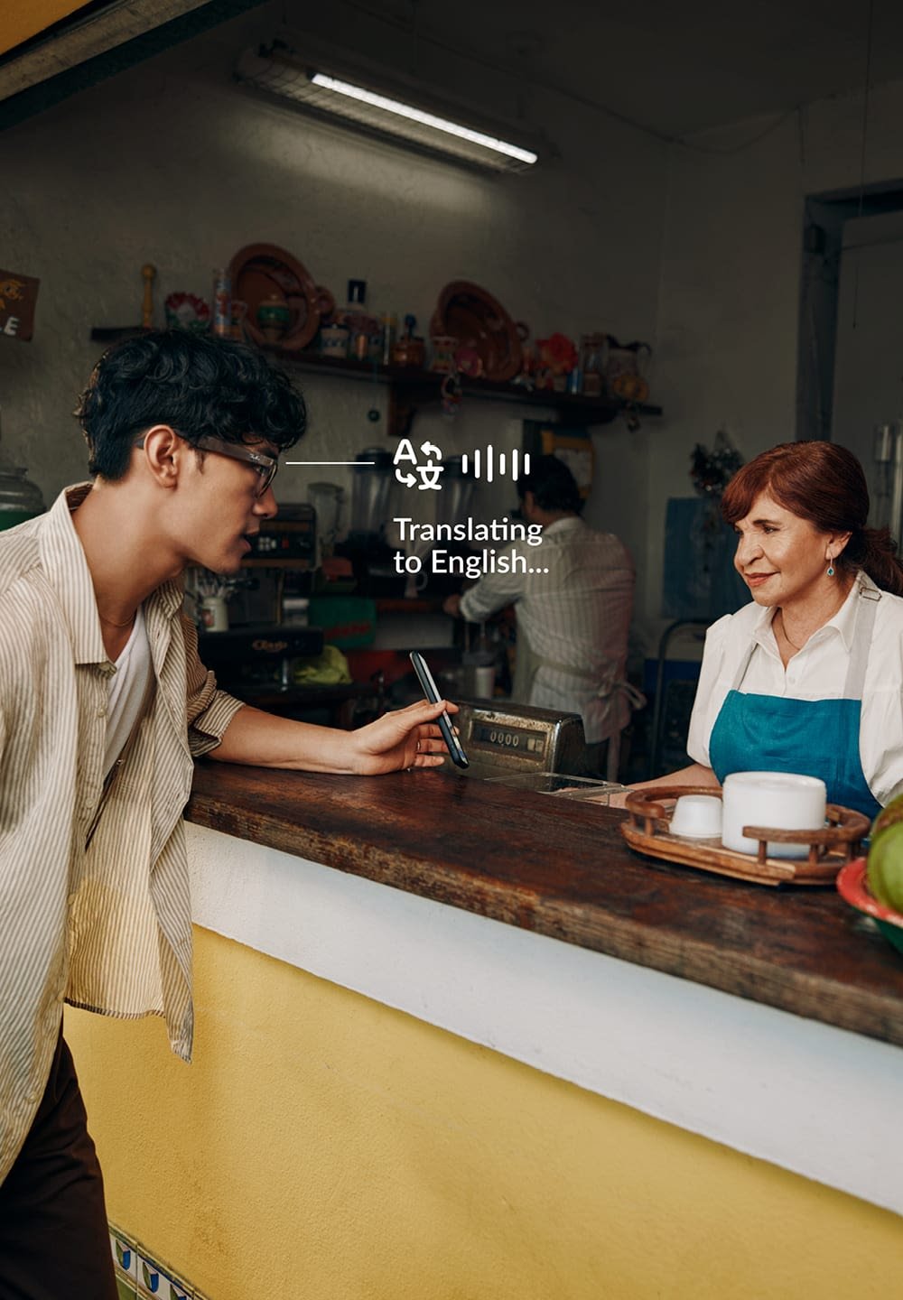 Comptoir avec une surface en bois, mur jaune et objets en céramique. Une personne en chemise rayée interagit avec une femme en tablier.