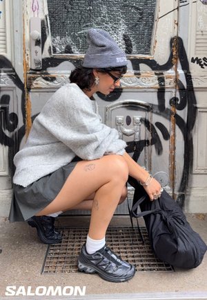 Woman in gray beanie, sweater, skirt, and black sneakers crouching by graffiti-covered door, holding a black bag.