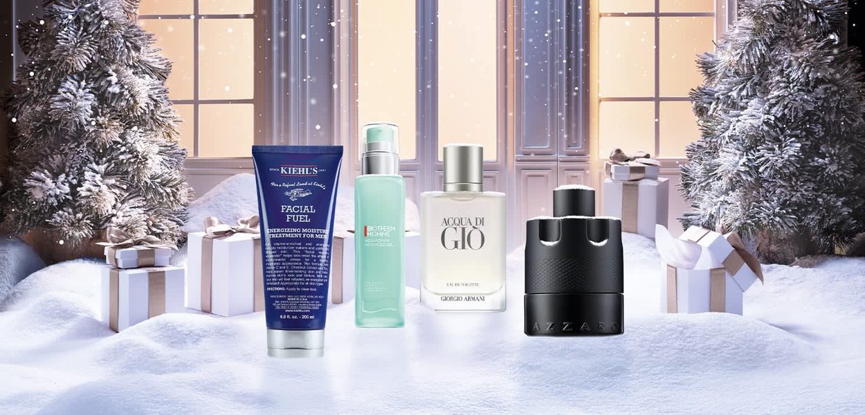 Satz von vier Pflegeprodukten vor einem verschneiten Hintergrund, darunter eine blaue Tube, eine grüne Flasche, ein klarer Parfümflakon und eine schwarze Flasche mit markanter Form.