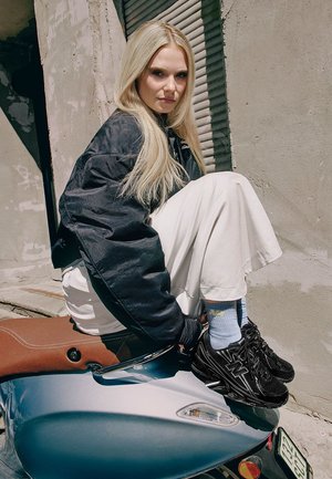 Black bomber jacket over white wide-legged trousers, paired with black New Balance sneakers and light blue socks, seated on a motorcycle.
