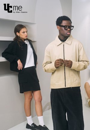 Béžová bunda na zips s golierom, nosená cez čierny rolák, skombinovaná s čiernymi kordovým nohavicami. Model v čiernom oblečení sa opiera v blízkosti.