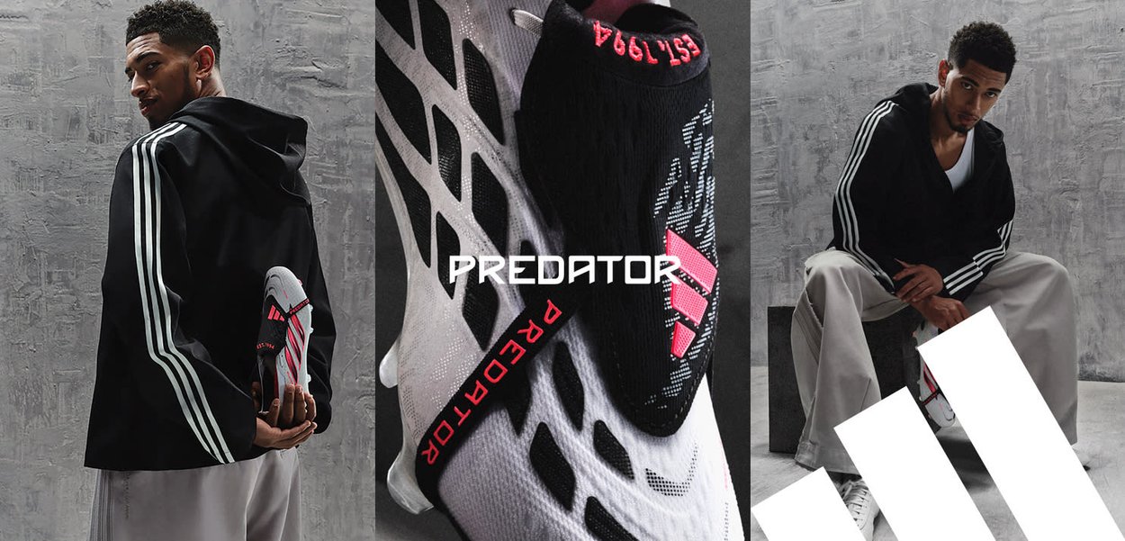 Mand iført en sort Adidas jakke med hvide striber holder en sort og hvid Adidas Predator fodboldstøvle med pink detaljer mod en grå baggrund.