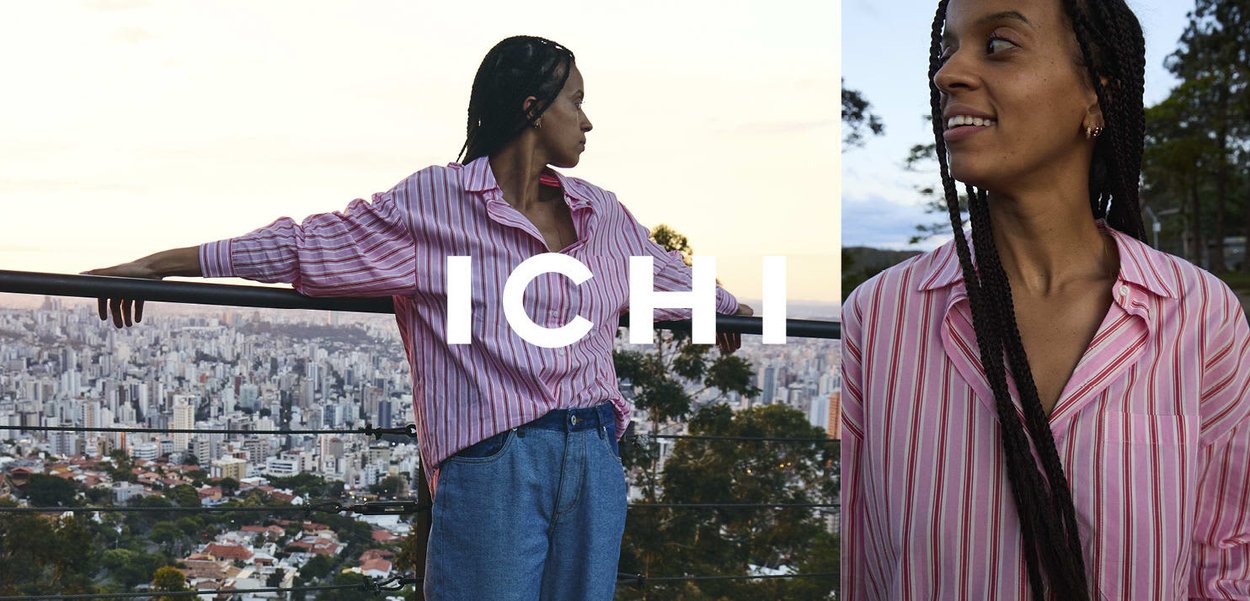 Frau mit geflochtenem Haar, die ein rosa gestreiftes Hemd und blaue Jeans trägt, steht an einem Glasgeländer und blickt bei Sonnenuntergang auf eine Stadtlandschaft.