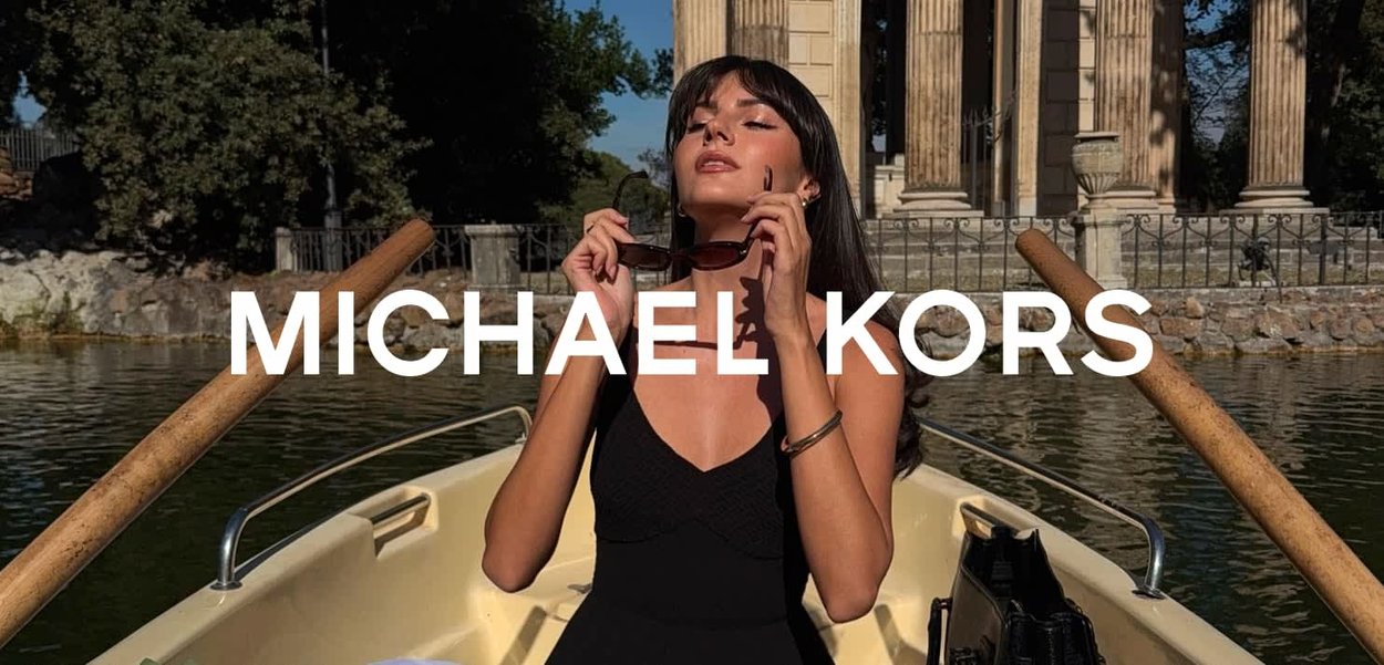 Abito nero lavorato a maglia, modella che tiene occhiali da sole, barca sull'acqua, strutture in pietra sullo sfondo, alberi verdi. Brand Michael Kors in evidenza.