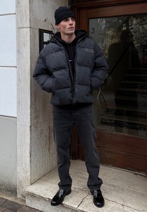 Giacca imbottita grigio scuro, felpa nera con cappuccio sotto, abbinata a jeans neri aderenti e scarpe eleganti nere lucide, ambientata davanti all'ingresso di un edificio.