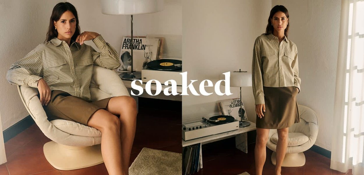 Model sitzt in einem cremefarbenen Stuhl, trägt ein kariertes grünes Hemd und einen braunen Rock. Im Hintergrund sind ein Plattenspieler, Schallplatten und natürliches Licht zu sehen.