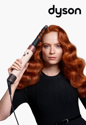 Donna con lunghi capelli rossi che utilizza un arricciacapelli Dyson per pettinarsi, indossando un top nero con una cintura.