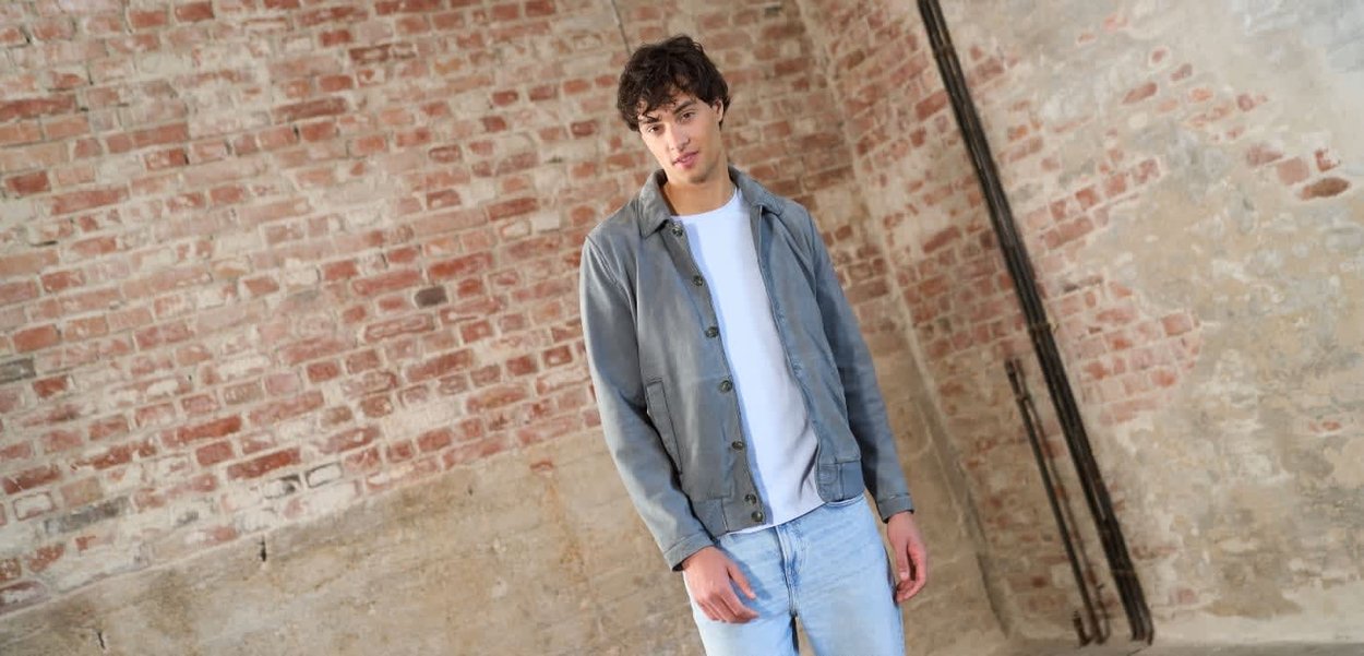 Giovane uomo in piedi contro un muro consumato di mattoni e cemento, indossa una giacca grigia, una maglietta bianca e jeans azzurri in una posa casual.