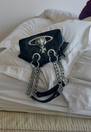 Sac à main en faux cuir noir avec des accents en chaîne argentée, un design structuré et un détail métallique circulaire sur le rabat. Placé sur une literie blanche.