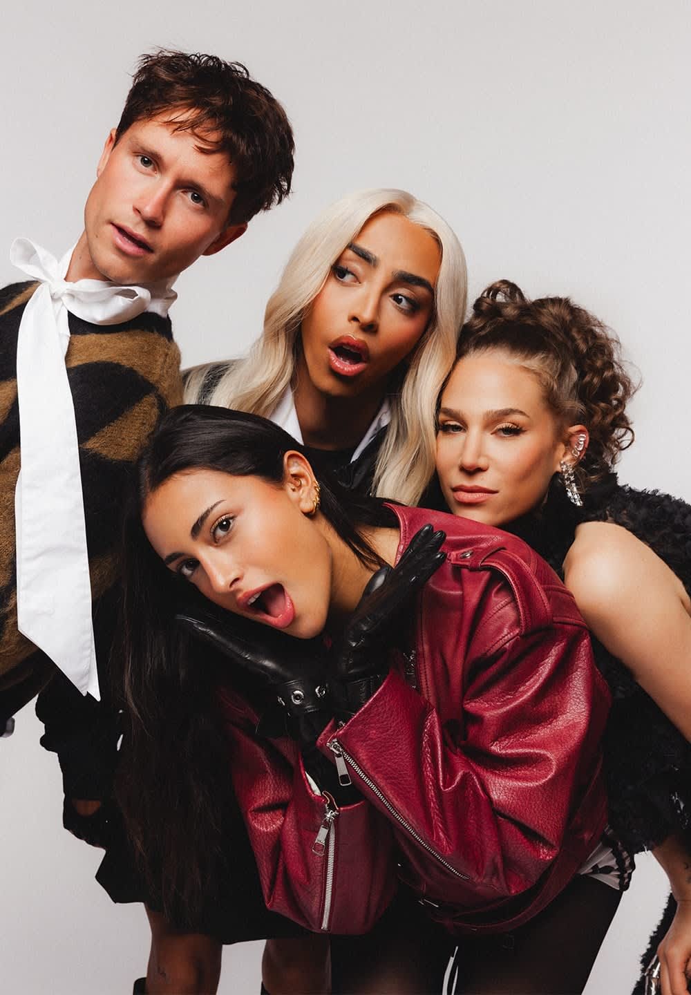 Quatre personnes posent côte à côte. Elles portent un mélange de vêtements texturés : un pull à motifs brun et noir, une chemise blanche, une veste en cuir bordeaux et un haut en fourrure noire.