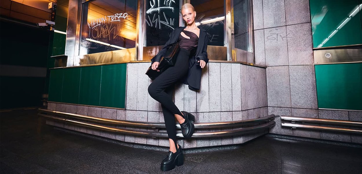 Donna bionda in abbigliamento nero e scarpe con platform appoggiata a un muro coperto di graffiti in un'ambientazione notturna urbana.