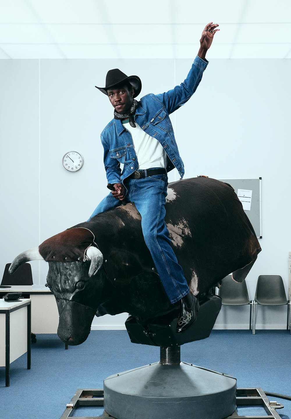 En rodeo-okse statue med en sort teksturert kropp og hvite detaljer; en person i blå denim, hvit skjorte og svart hatt poserer oppå den.