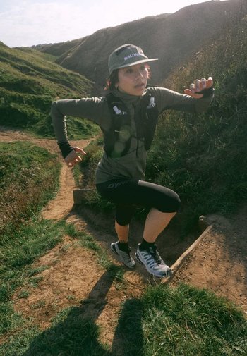 Echipament de alergare, inclusiv un tricou lung verde, colanți negri, o șapcă gri cu text și adidași gri pentru alergare. Fundal cu un traseu montan.