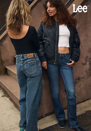 To kvinner snakker sammen i trapper. Den ene har på seg en svart tettsittende topp og vide blå jeans med et Lee-merke; den andre har på seg en kort hvit topp og mørke jeans.