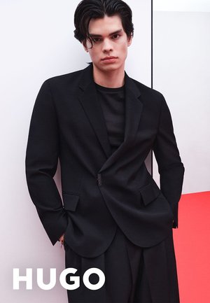Schwarzer maßgeschneiderter Blazer mit Reverskragen und Fronttasche. Getragen über einem eng anliegenden schwarzen Hemd. Individuum posiert vor einem minimalistischen Hintergrund.