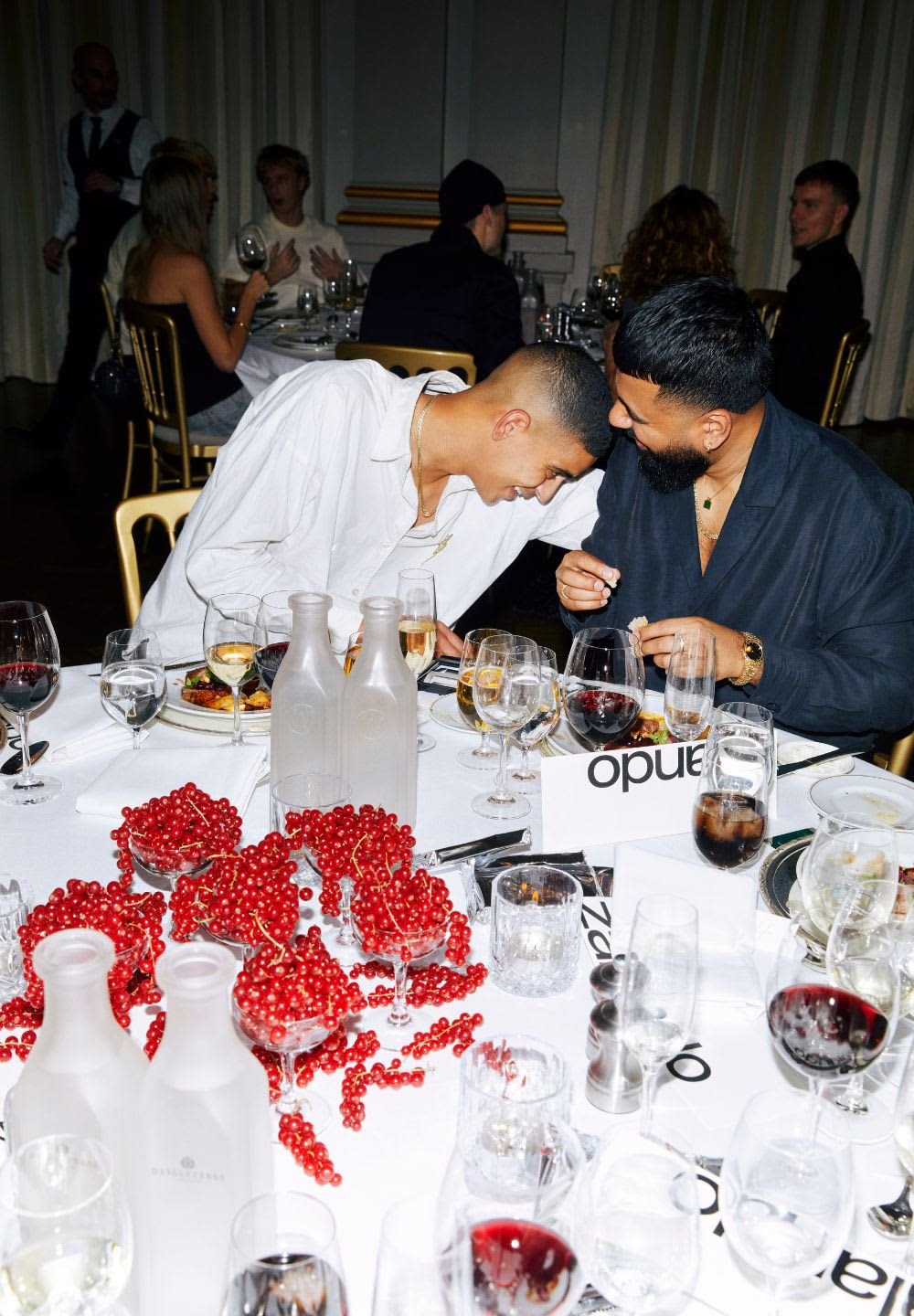 Bord dækket med forskellige glas, to klare flasker, et dekorativt arrangement af klare røde bær og tallerkener med mad i en spisesituation.