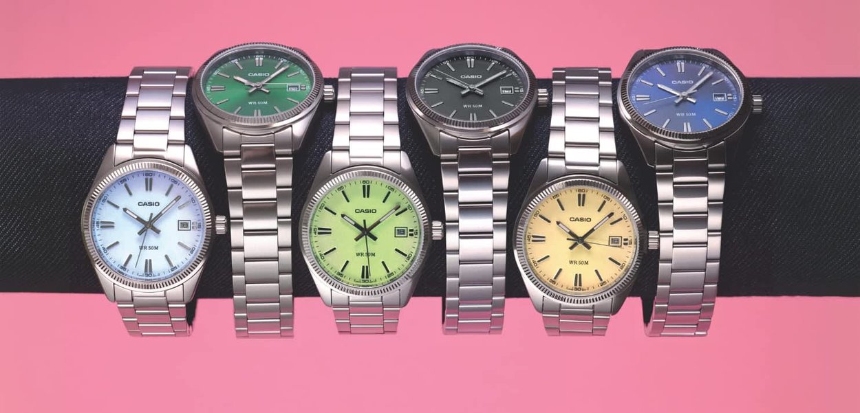 Seis relojes de pulsera Casio con correas de metal plateado y esferas de diferentes colores expuestos sobre una barra negra sobre un fondo rosa.