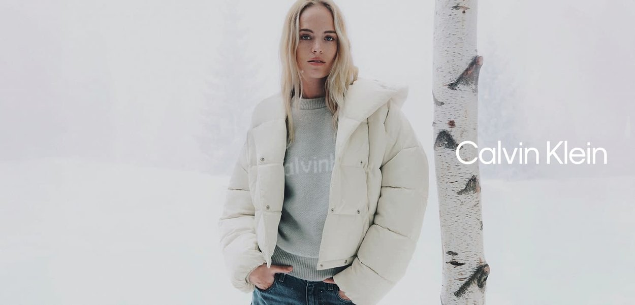 Chaqueta blanca acolchada con capucha, con textura acolchada y botones a presión, llevada sobre un suéter gris con el nombre de la marca.