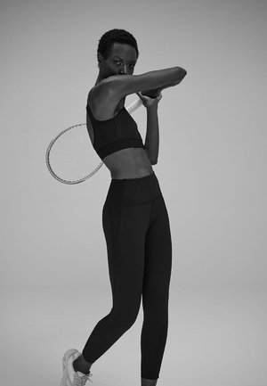 Top corto sportivo nero e leggings a vita alta, texture liscia, abbinati a una racchetta da tennis in un contesto monocromatico.