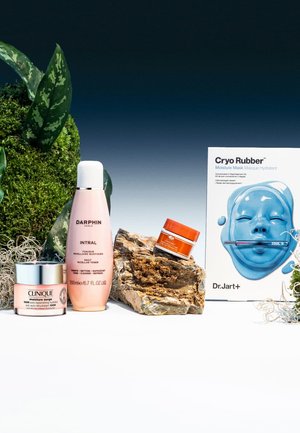 Three skincare products: a pink micellar toner in an opaque bottle, a white cream in a silver-lidded jar, and an orange cream in a small jar, alongside a blue mask box. Rich green foliage and textured rocks complete the arrangement.