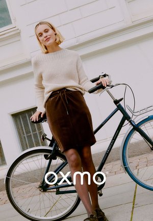 Un pull en laine beige, une jupe en corduroy marron, se tenant à côté d'un vélo noir avec des accents bleus. Pavé lisse, bâtiment de couleur claire en arrière-plan.