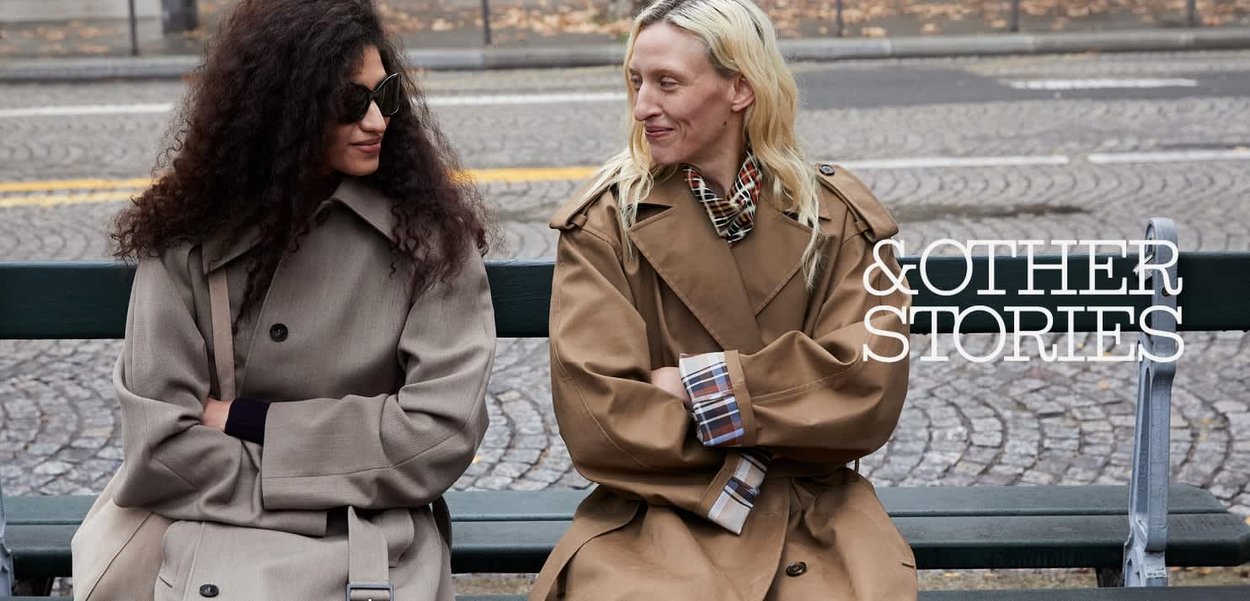 To kvinder iført beige trenchcoats sidder på en bænk udendørs, ser på hinanden og smiler, med en brostensbelagt gade i baggrunden.