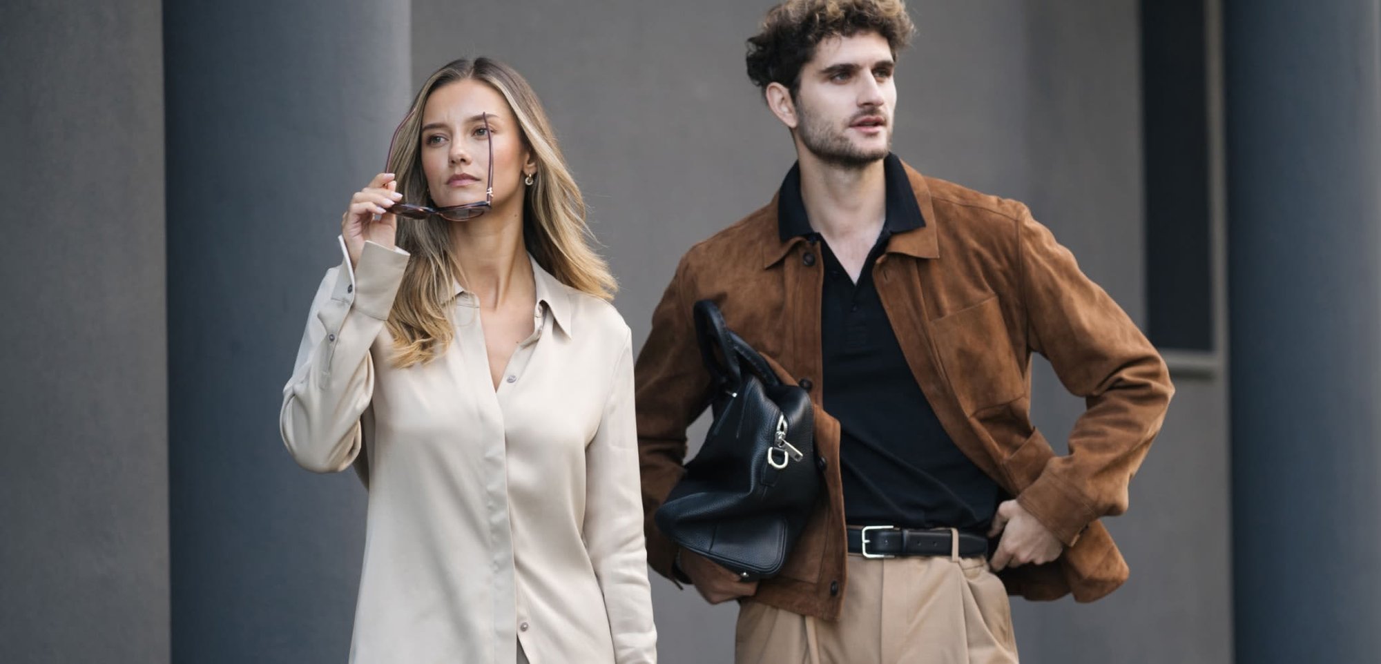A woman in a beige satin button-up and trousers holds sunglasses; a man in a brown suede jacket and black polo carries a black bag.