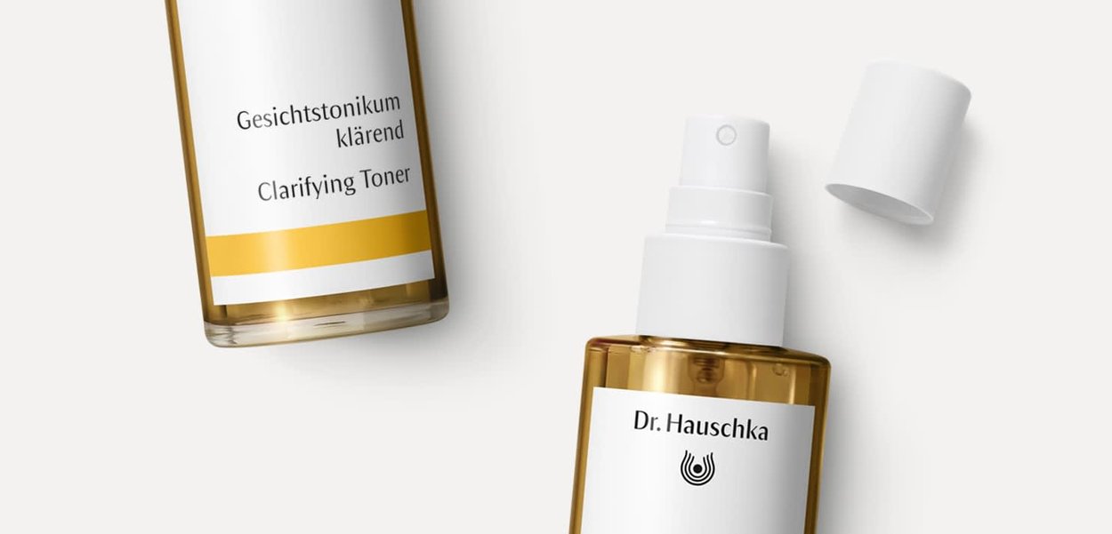 Dr. Hauschka Klärendes Gesichtswasser in einer klaren, bernsteinfarbenen Flasche mit einem weißen Sprühaufsatz. Das Etikett zeigt schwarzen Text und einen gelben horizontalen Streifen.