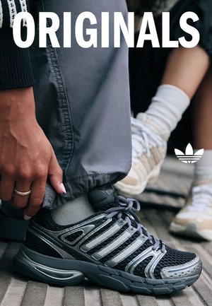 Chaussures de sport noires et grises avec tige en mesh, accents métalliques et système de laçage. Complétées par un pantalon de jogging gris et des chaussettes claires.