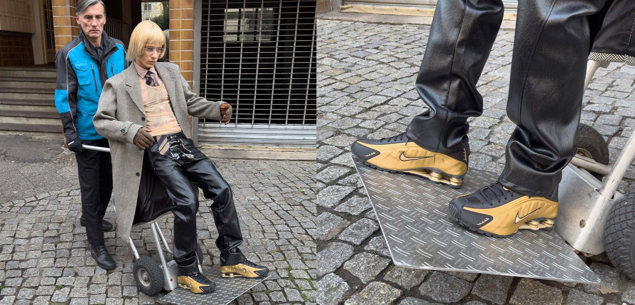 Eine Schaufensterpuppe in einem maßgeschneiderten grauen Mantel und transparentem Hemd, sitzend auf einem rollbaren Wagen. Schwarze Ledershosen, goldakzentuierte Schuhe, urbanes Umfeld.