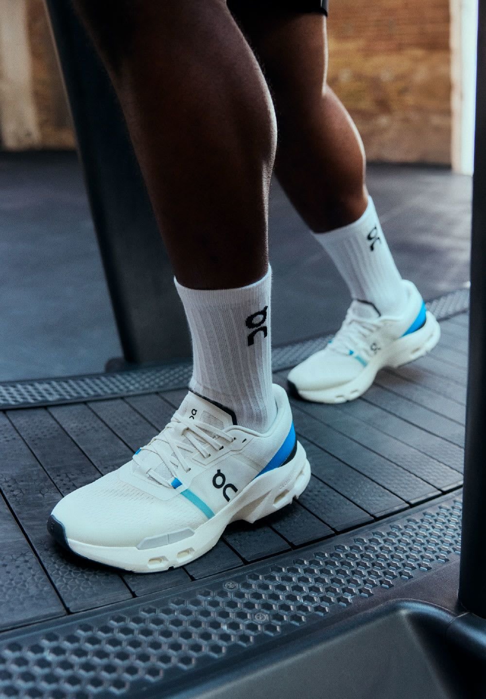 Weiße Sportschuhe mit blauen Akzenten, gepolsterten Sohlen und Logo an der Seite, kombiniert mit weißen Socken, auf einem Laufband in einer Fitnessumgebung.