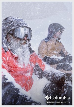 Zwei Personen sitzen im tiefen Schnee, eine in einer roten Jacke mit fellgefütterter Kapuze und einer Schutzbrille, die andere in einer braunen Jacke, beide mit Schneeflocken bedeckt.