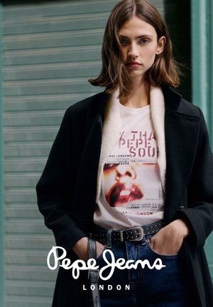 Jonge vrouw die een zwarte jas draagt, een wit T-shirt met een lippenafdruk en blauwe jeans met een zwarte riem, staand tegen een groene luifel.