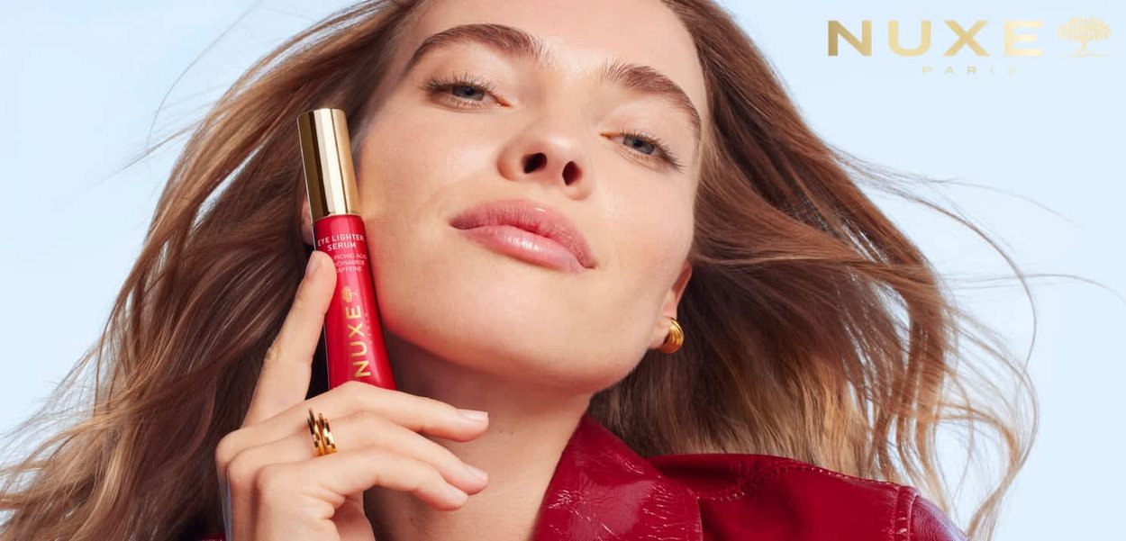 Donna con capelli lunghi tiene vicino al viso il siero illuminante per occhi rosso NUXE, indossa una giacca rossa, orecchini d'oro e un anello, su sfondo chiaro.