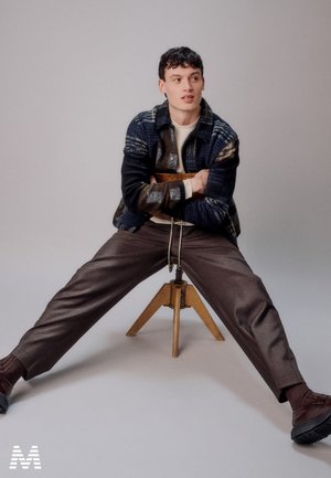 Veste en tartan bleu marine et gris, pull crème, pantalon marron, assis sur un tabouret en bois trépied, avec une posture détendue et un fond neutre.