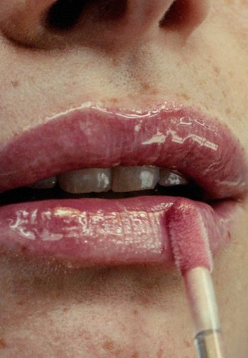 Applicatore di gloss per labbra utilizzato su labbra piene, lucide, di colore rosa pallido con una texture liscia e una lucentezza naturale, evidenziata da lievi imperfezioni.