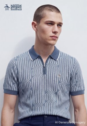Jonge man met kort haar draagt een blauw, patroonrijk zip-up polohemd met een pinguïnlogo op de borst, staand tegen een effen lichte achtergrond.
