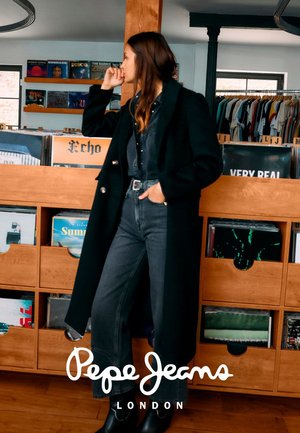 Schwarzer langer Mantel über einem dunkelblauen Jeanshemd und hoch taillierten Jeans. Model mit lockigem Haar steht neben Holzregalen mit Vinyl-Schallplatten.