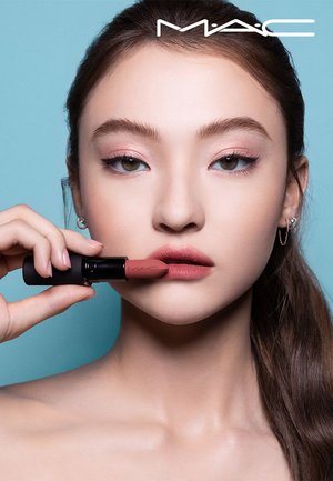 Jonge vrouw met roze oogschaduw brengt gedempte rozenkleurige lippenstift aan, terwijl ze MAC-lippenstift dicht bij haar lippen houdt tegen een lichtblauwe achtergrond.