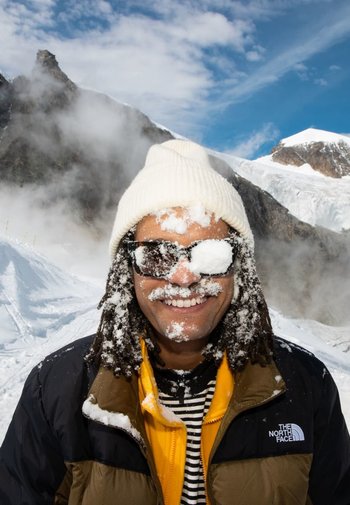 Person mit langen, dunklen Haaren, die eine weiße Strickmütze und Sonnenbrille trägt, bedeckt mit Schnee, gekleidet in eine schwarz-gelbe Jacke, vor einer verschneiten Landschaft.