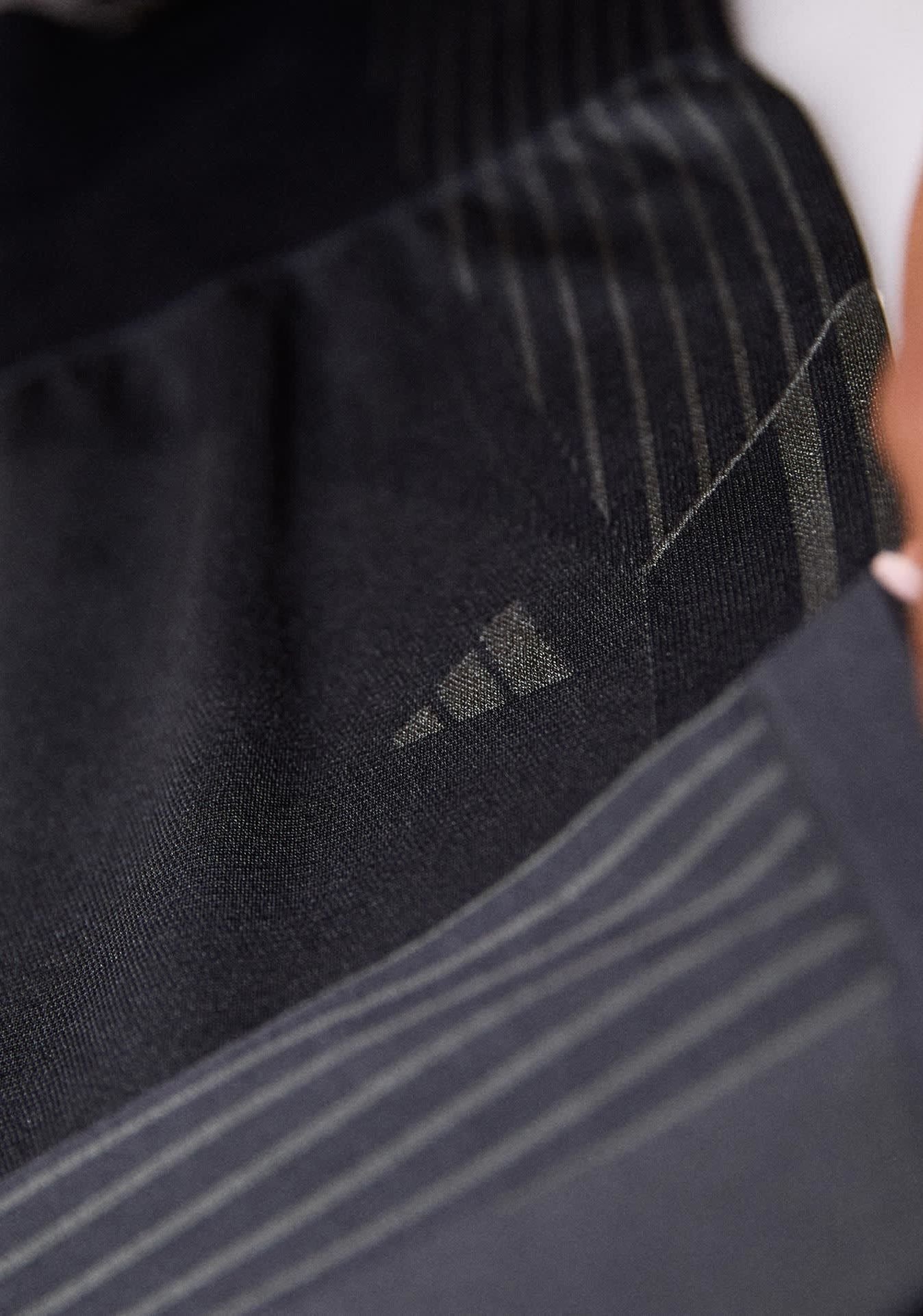 Schwarzer sportlicher Stoff mit dezenten vertikalen Streifen und einer strukturierten Oberfläche. Enthält ein kleines Adidas-Logo in einem dunkleren Farbton auf dem Stoff.